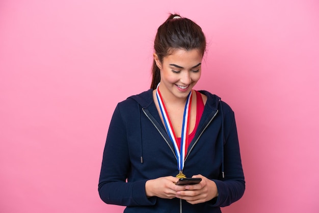 Młoda brazylijska sportowa kobieta z medalami na białym tle na różowym tle wysyła wiadomość za pomocą telefonu komórkowego