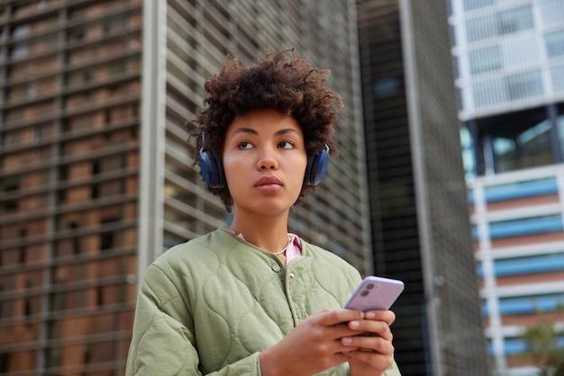 Młoda brunetka cieszy się muzyką ze słuchawkami trzyma telefon komórkowy pobiera piosenkę na playlistę ma na sobie kurtkę