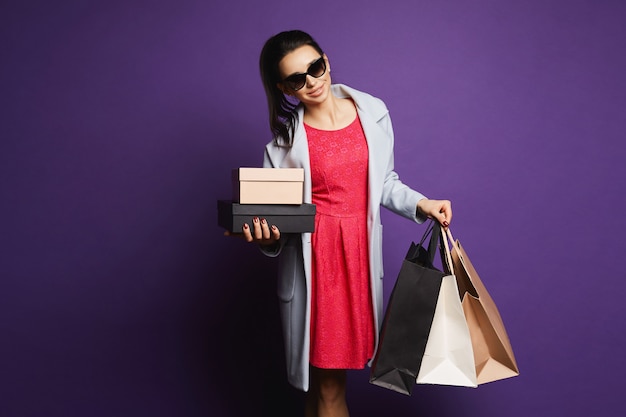 Młoda brunetka dama w płaszcz, czerwona sukienka i okulary przeciwsłoneczne z torby na zakupy w jej ręce