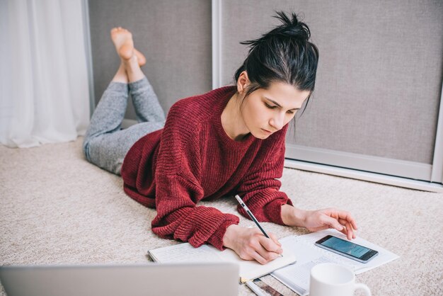 Młoda brunetka gospodyni domowa leżąca na podłodze za pomocą laptopa zarządzającego budżetem krajowym robienie notatek ołówkiem obliczanie na papierze i dokumentach smartfona Biznesowa kobieta pracująca w domu Styl życia