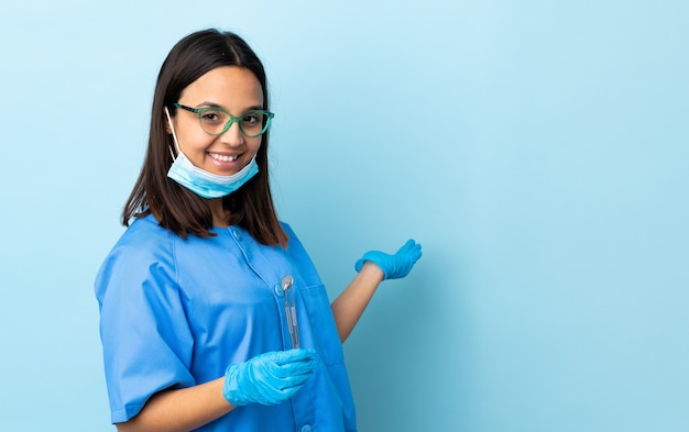 Młoda brunetka kobieta dentysta rasy mieszanej, trzymając narzędzia na izolowanych wyciągając ręce na bok, aby zachęcić do przyjazdu