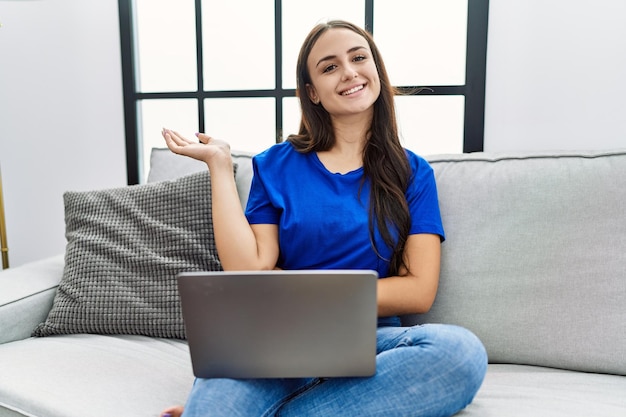 Młoda brunetka kobieta korzystająca z laptopa w domu uśmiecha się wesoło, przedstawiając i wskazując dłonią patrząc w kamerę.