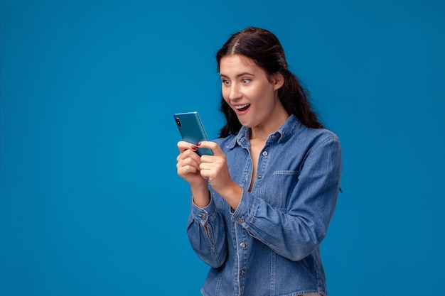 Młoda brunetka kobieta pozuje ze smartfonem stojącym na niebieskim tle.