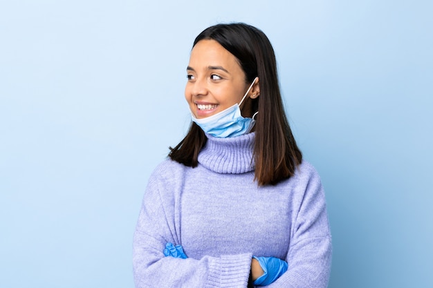 Młoda brunetka kobieta rasy mieszanej ochrony przed koronawirusem z maską i rękawice na pojedyncze niebieskie ściany patrząc po stronie