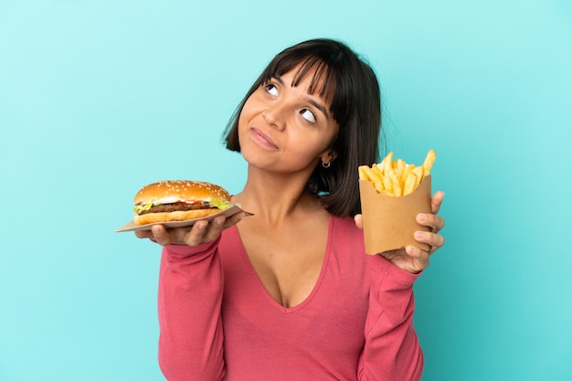 Młoda brunetka kobieta trzyma burgera i smażone frytki na odosobnionym niebieskim tle