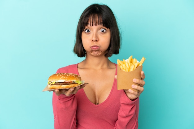 Młoda brunetka kobieta trzyma burgera i smażone frytki na odosobnionym niebieskim tle