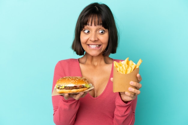 Młoda brunetka kobieta trzyma burgera i smażone frytki nad odosobnioną niebieską ścianą