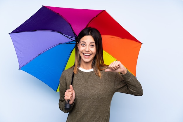 Młoda brunetka kobieta trzymając parasol nad niebieską ścianą dumny i zadowolony