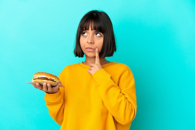 Młoda brunetka kobieta trzymająca burgera na białym tle, mająca wątpliwości, patrząc w górę