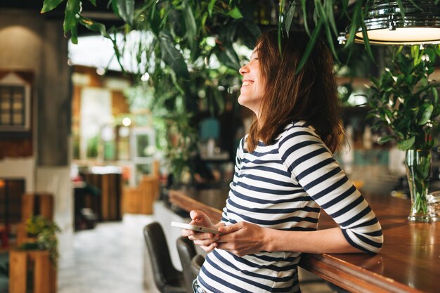 Młoda brunetka kobieta ubrana w pasiasty longsleeve z telefonem komórkowym w rękach w zielonym eco cafe city life