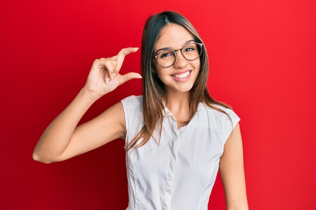 Młoda brunetka kobieta ubrana w zwykłe ubrania i okulary, uśmiechnięta i pewna siebie, gestykulująca ręką robiąca mały znak z palcami patrzącymi i aparatem. koncepcja środka.