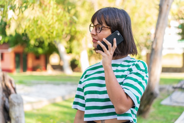 Młoda brunetka kobieta w parku prowadzi z kimś rozmowę przez telefon komórkowy