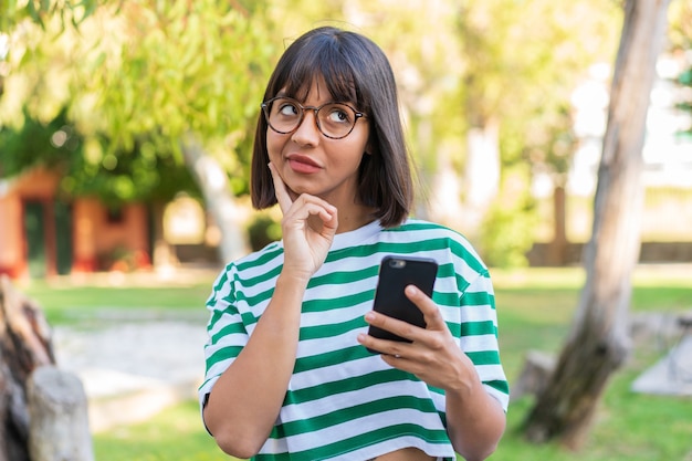Młoda brunetka kobieta w parku przy użyciu telefonu komórkowego i myślenia