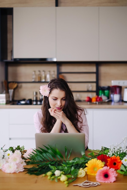 Młoda brunetka kobieta z długimi falującymi włosami pracuje na laptopie z kwiatami na biurku z kuchnią w tle Utalentowana kwiaciarnia rozwija sprzedaż online przygotowuje się do warsztatów