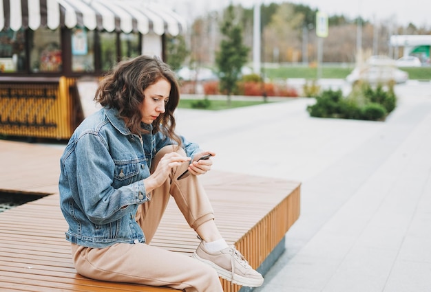 Młoda brunetka kobieta z kręconymi włosami w swobodnym ubraniu, używająca telefonu komórkowego na ulicy