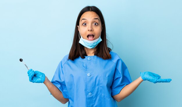 Młoda brunetka mieszająca biegowa dentysta kobiety mienia narzędzia nad ścianą z szokującym wyrazem twarzy