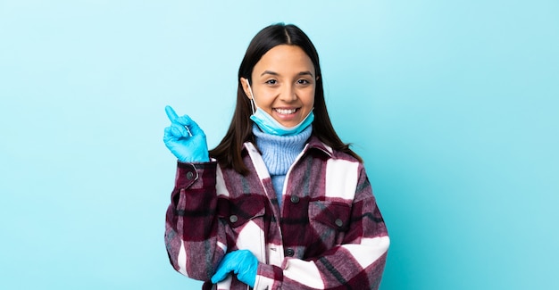 Młoda brunetka mieszająca biegowa kobieta ochrania przed koronawirusem z maską i rękawiczkami nad odosobnioną błękit ścianą szczęśliwą i wskazuje up