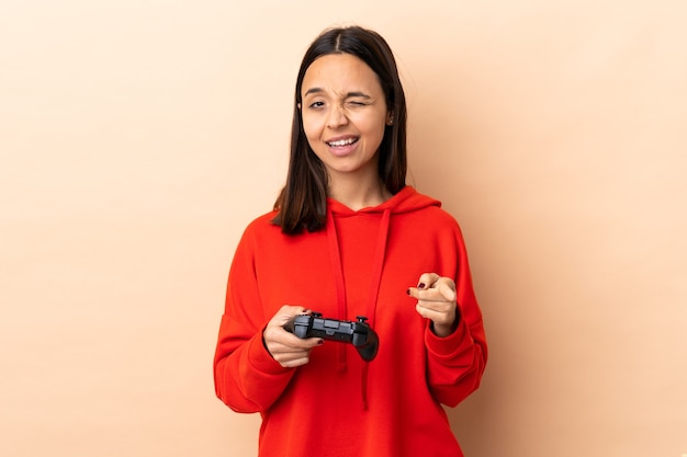 Młoda brunetka mieszanej rasy kobieta bawi się kontrolerem gier wideo na odosobnionej ścianie wskazuje palcem na ciebie