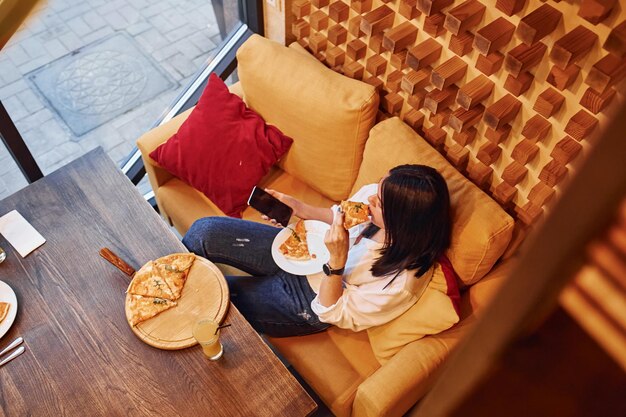 Młoda brunetka siedzi w pomieszczeniu z pizzą i smartfonem w ręku.