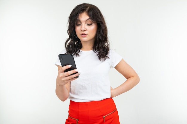 Młoda brunetka w białej koszulce z powątpiewaniem patrzy na smartfona, trzymając go w dłoni na białym tle odizolowane z miejsca na kopię.
