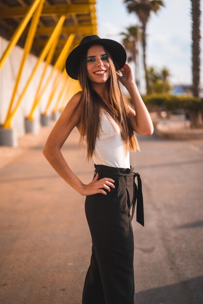 Młoda brunetka w czarnym kapeluszu i spodniach na żółtej promenadzie