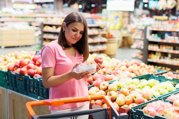 Młoda brunetka wybiera owoce na ladzie w supermarkecie Gospodyni domowa robi zakupy na rynku, stojąc w pobliżu sklepu warzywnego z wózkiem spożywczym