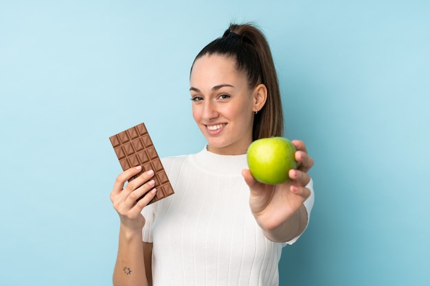Młoda brunetki kobieta nad odosobnioną błękit ścianą bierze czekoladową pastylkę w jednej ręce i jabłko w drugiej