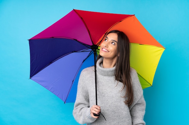 Młoda brunetki kobieta trzyma parasol nad odosobnioną błękit ścianą śmia się i przygląda up