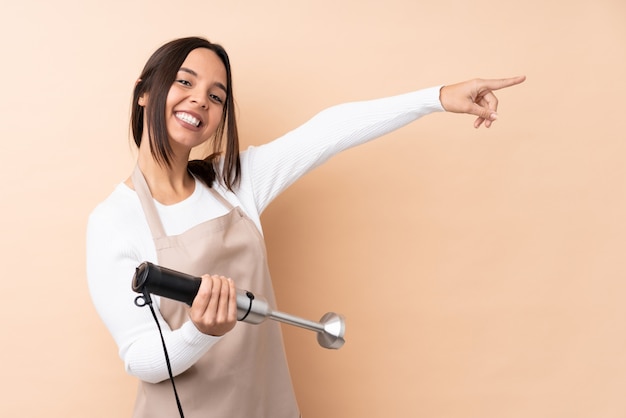 Młoda brunetki kobieta używa ręka blender nad odosobnionym ściennym wskazuje palcem strona i przedstawia produkt