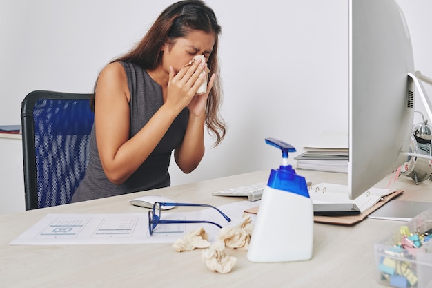 Młoda businesswoman kichanie w bibułkę, gdy siedzisz przy biurku, grypa i koncepcja alergii