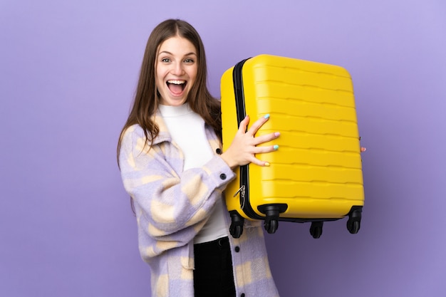 Młoda caucasian kobieta na purpury ścianie w wakacje z podróży walizką i zaskakująca
