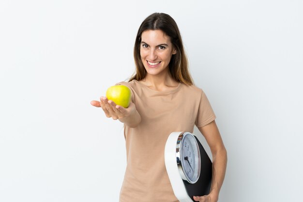 Młoda caucasian kobieta trzyma biel waży maszynę i oferuje jabłka na biel ściany