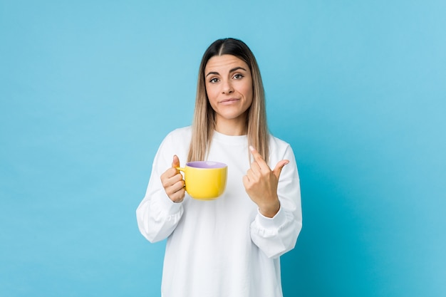 Młoda caucasian kobieta trzyma filiżankę kawy wskazuje z tobą palcem, jakby zapraszający przychodził bliżej.