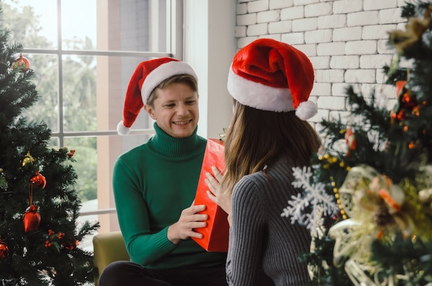 Młoda Caucasian Para Z Czerwonym Santa Kapeluszowym Zaskakiwaniem Daje Boże Narodzenie Prezenta Pudełku Dziewczyna Z Choinki świętowaniem W Domu