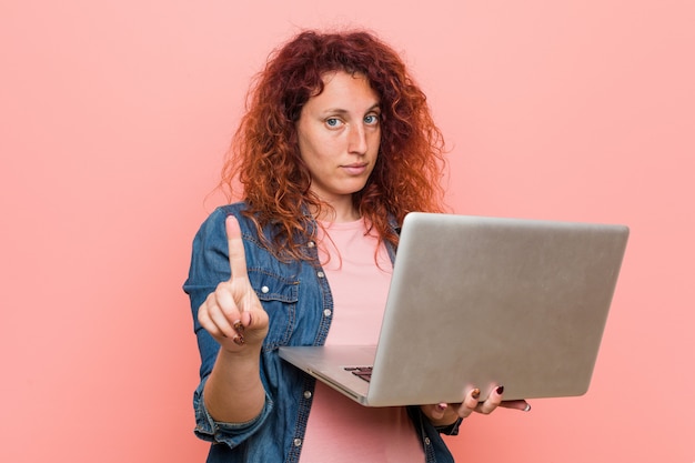 Młoda caucasian rudzielec kobieta trzyma laptop pokazuje liczbę jeden z palcem.