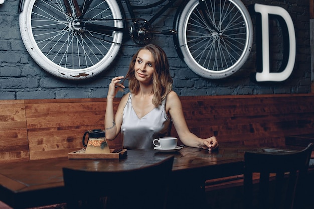 Młoda charyzmatyczna zalotna blondynka siedzi w sztuce kawy w stylu retro na tle ceglanego muru i wiszącego roweru. Koncepcja tematyczna