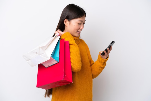 Młoda Chinka na białym tle trzyma torby na zakupy i pisze wiadomość telefonem komórkowym do przyjaciela