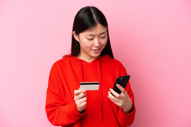 Młoda Chinka odizolowana na różowym tle, kupująca za pomocą telefonu komórkowego za pomocą karty kredytowej