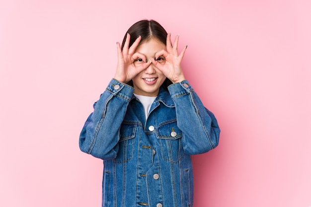 Młoda Chinka, pozowanie w różowej ścianie na białym tle pokazuje znak porządku na oczy