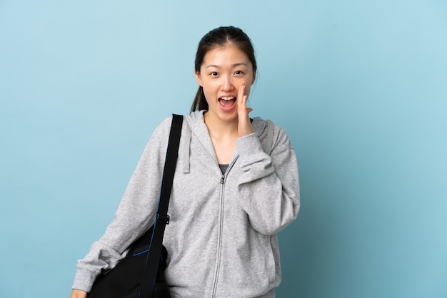 Młoda Chinka sportowa z torbą sportową na białym tle niebieski krzyczy z szeroko otwartymi ustami