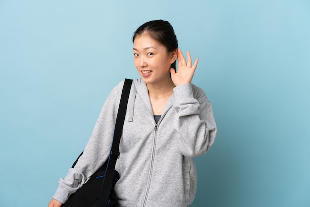 Młoda Chinka sportowa z torbą sportową na białym tle niebieski słuchając czegoś, kładąc rękę na uchu