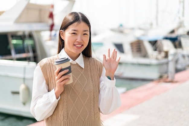 Młoda Chinka Trzyma Kawę Na Wynos Na Zewnątrz Pozdrawiając Ręką Z Radosnym Wyrazem Twarzy