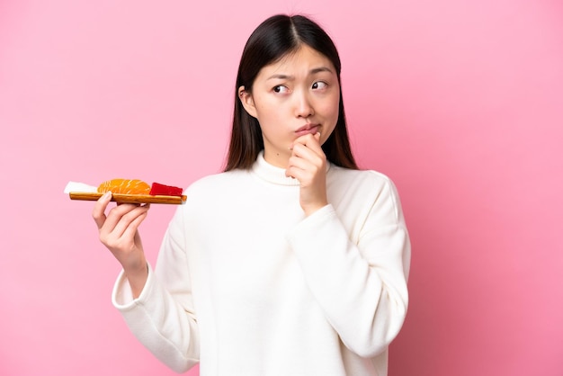 Młoda Chinka trzymająca sashimi odizolowana na różowym tle, mająca wątpliwości i myślenie