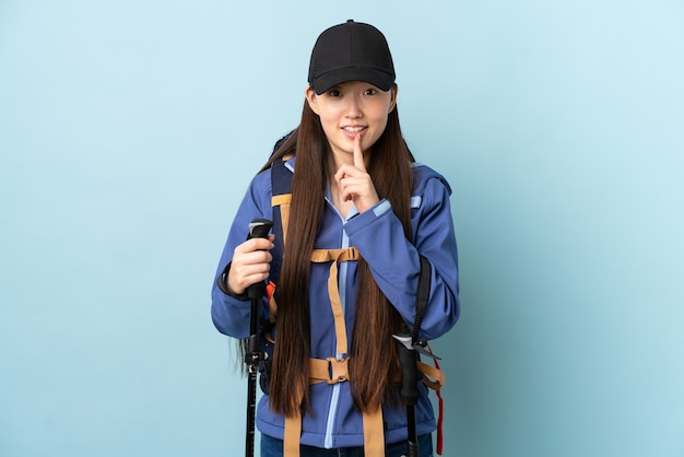 Młoda Chinka z plecakiem i kijkami trekkingowymi na białym tle niebieski pokazując znak gestu ciszy wkładając palec w usta