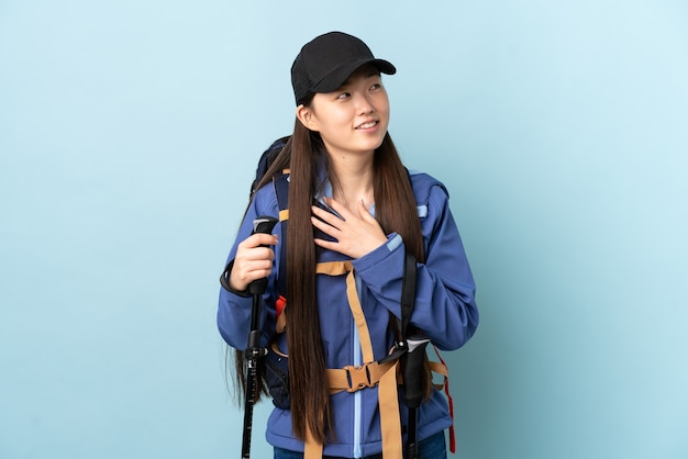 Młoda chińska dziewczyna z plecakiem i kijkami trekkingowymi nad izolowaną niebieską ścianą patrząc w górę podczas uśmiechu