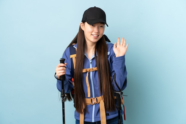 Młoda chińska dziewczyna z plecakiem i kijkami trekkingowymi nad izolowaną niebieską ścianą salutując ręką z happy wypowiedzi