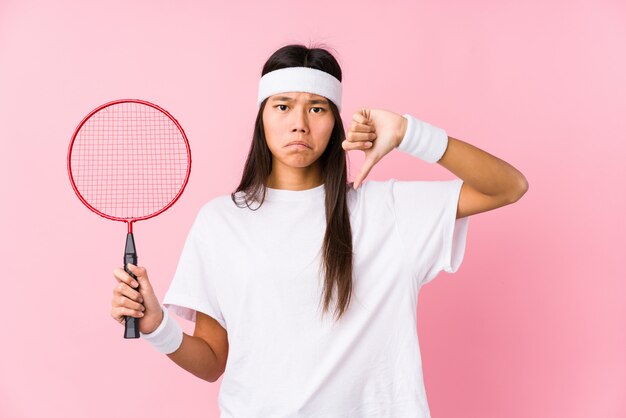 Młoda chińska kobieta bawić się badminton w różowej ścianie pokazuje niechęć gest, kciuki zestrzela. Pojęcie nieporozumienia.