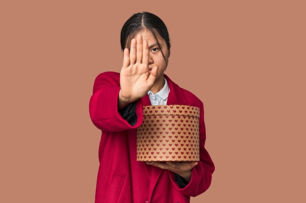 Młoda chińska kobieta trzymająca pudełko pełne serca stojąca z wyciągniętą ręką pokazującą znak zatrzymania