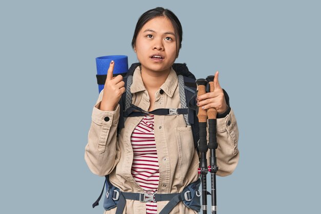 Młoda chińska kobieta z sprzętem turystycznym gotowa na przygodę wskazująca do góry nogami z otwartymi ustami
