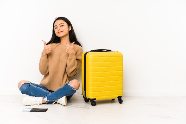 Młoda chińska podróżniczka siedzi na podłodze z izolowaną walizką, podnosząc kciuki do góry, uśmiechnięta i pewna siebie.